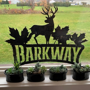 May include: Black metal sign with a deer silhouette and maple leaves, reading "BARKWAY". Four black pots with green succulents are arranged on a windowsill.