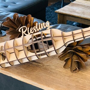 May include: A wooden wine bottle holder with a cutout design. The holder is labeled "Riesling" and is sitting on a wooden surface.