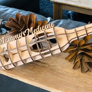 May include: A wooden wine bottle holder with the words "Mommy's Medicine" carved into the side. The holder is made of light brown wood and has a grid pattern. The holder is sitting on a wooden surface.