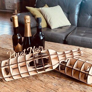 May include: Wooden wine bottle holder with the words "Pinot Noir" carved into the side. Three wine bottles are displayed in the holder, one is a champagne bottle, one is a red wine bottle, and one is a clear bottle with a gold label.