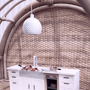 May include: A miniature white kitchen set with a white sink, a white oven with a brown door, and white cabinets with black handles. The kitchen is set against a woven wicker wall with a white pendant light hanging above.