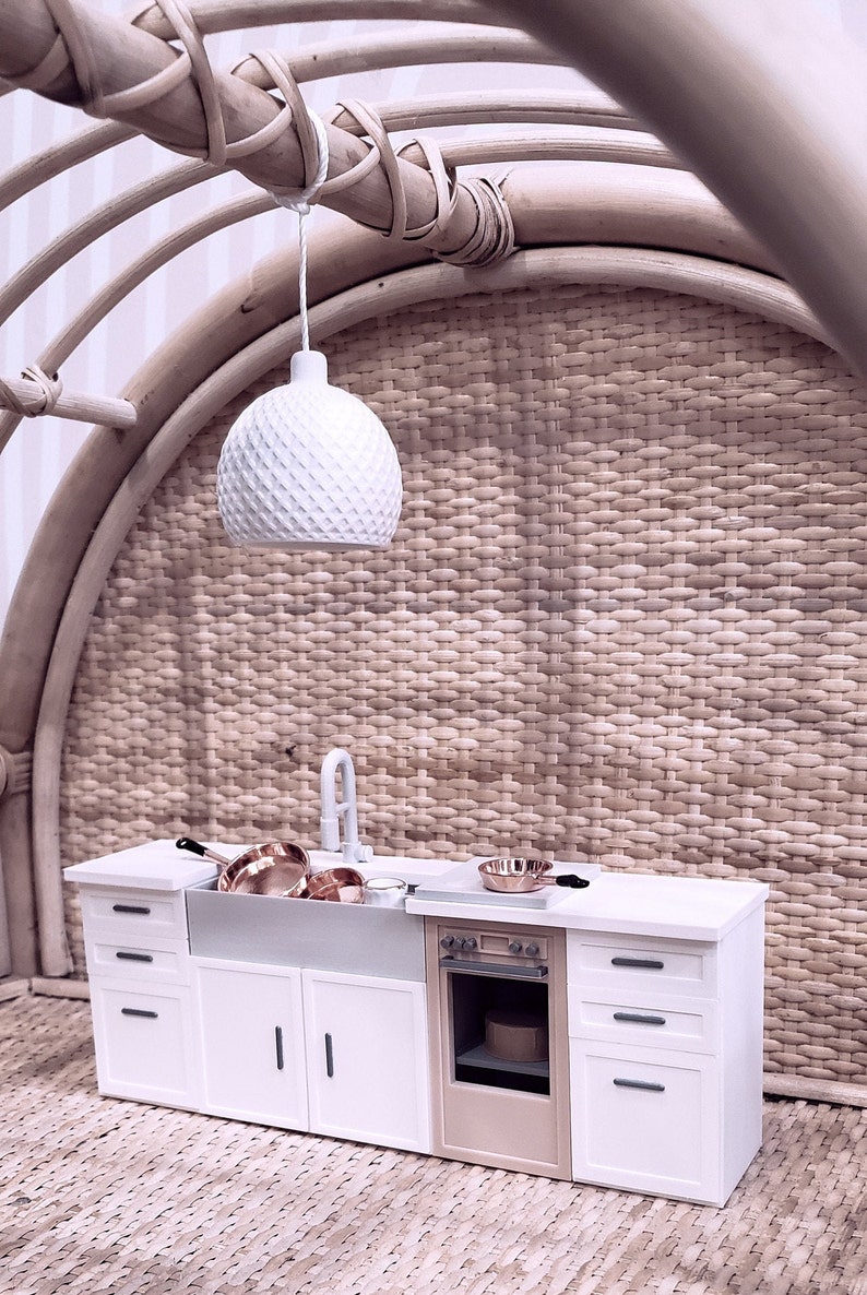 May include: A miniature white kitchen set with a white sink, a white oven with a brown door, and white cabinets with black handles. The kitchen is set against a woven wicker wall with a white pendant light hanging above.