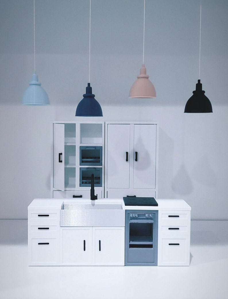 May include: A miniature white kitchen set with a stainless steel sink, a black stovetop, and a gray oven. The kitchen has white cabinets with black handles and three pendant lights hanging above. The lights are blue, pink, and black.