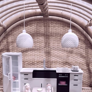 May include: A miniature dollhouse kitchen set with white cabinets, a black sink, and a white oven. Two small white pendant lights hang above the kitchen. Two small figurines of rabbits are sitting at a table with a white plate.