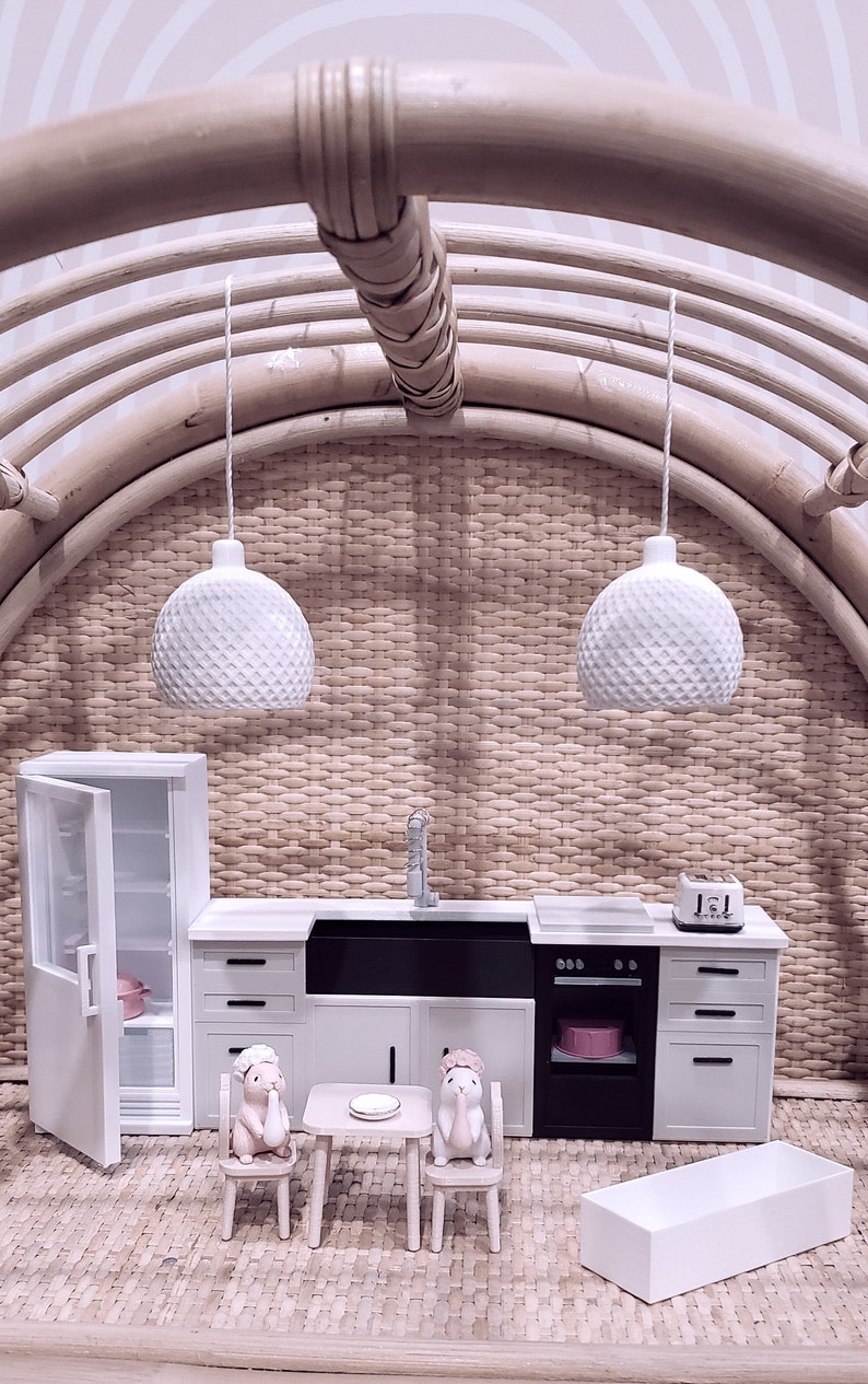 May include: A miniature dollhouse kitchen set with white cabinets, a black sink, and a white oven. Two small white pendant lights hang above the kitchen. Two small figurines of rabbits are sitting at a table with a white plate.