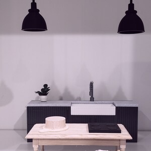 May include: A miniature kitchen set with a black countertop, a white sink, a wooden table, a wooden crate, and a woven basket. Two black pendant lights hang above the countertop.