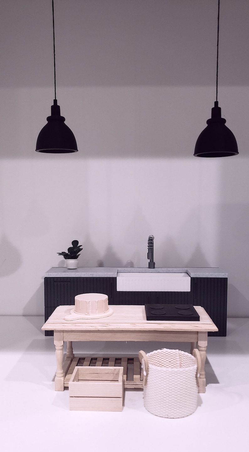 May include: A miniature kitchen set with a black countertop, a white sink, a wooden table, a wooden crate, and a woven basket. Two black pendant lights hang above the countertop.