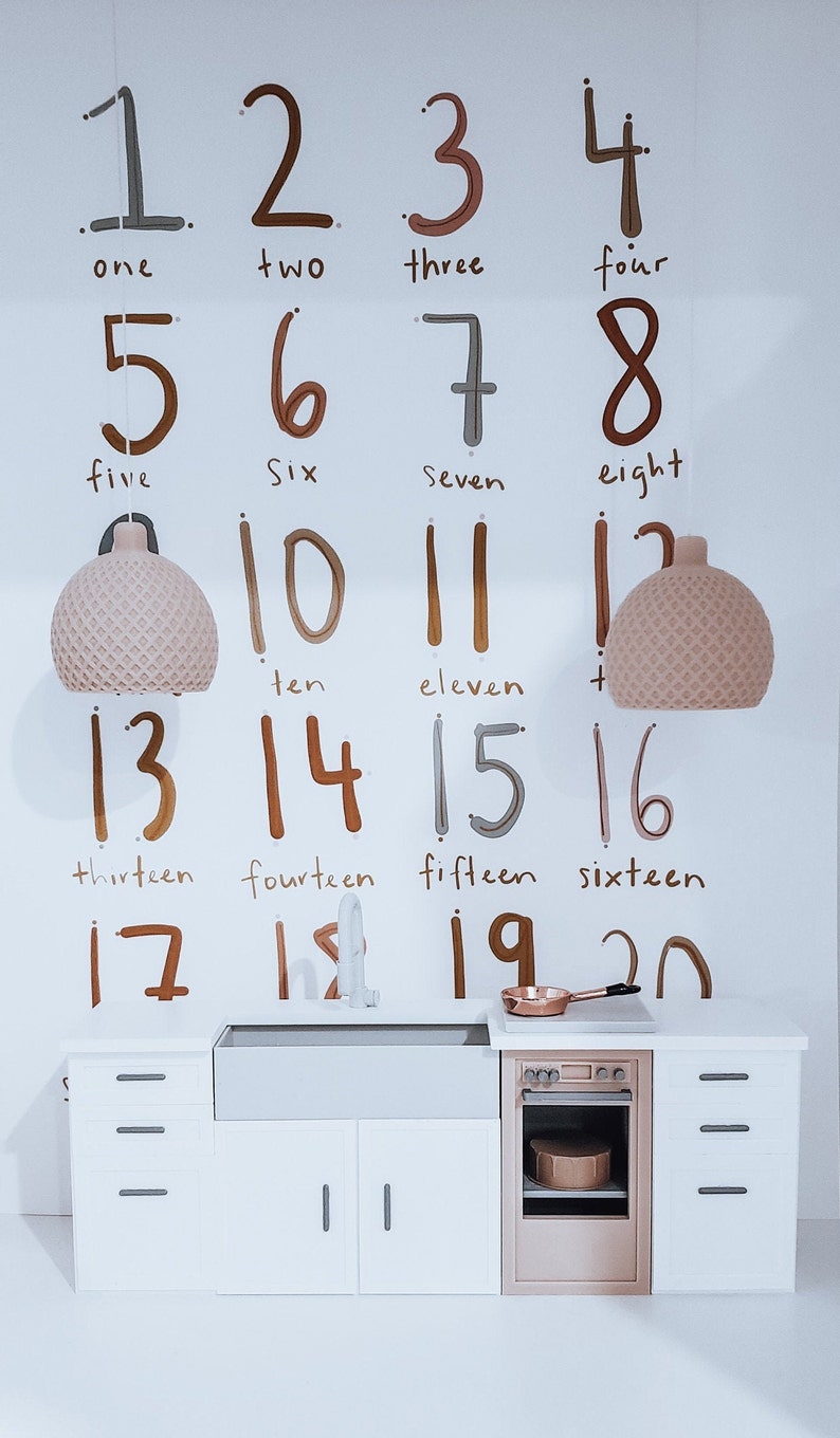 May include: A white wall with handwritten numbers from one to twenty, with the corresponding word for each number written below. There is a miniature kitchen set with a sink, oven, and cabinets below the numbers.