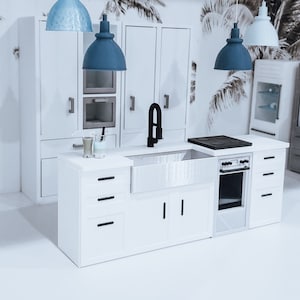 May include: A white kitchen island with a farmhouse sink, a black faucet, and a black stovetop. The island has three drawers and two cabinet doors. There are three pendant lights hanging above the island, two blue and one light blue.