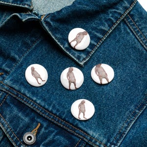 May include: Five white round buttons with images of a person in a white suit on a blue denim jacket.