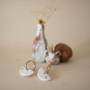 Op de afbeelding: Een paar witte keramische oorbellen met gouden ringen. De oorbellen hebben een halve cirkelvorm met kleine bruine spikkels. Op de achtergrond staan twee keramische vazen en een bruin object, alles op een beige oppervlak.