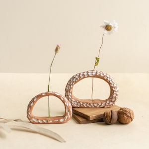 Pode incluir: Dois vasos de cerâmica cor de terracota com um design texturizado e salpicado de branco. Cada vaso contém uma única flor de haste. Os vasos são exibidos com blocos de madeira e nozes, criando uma estética natural e terrosa.