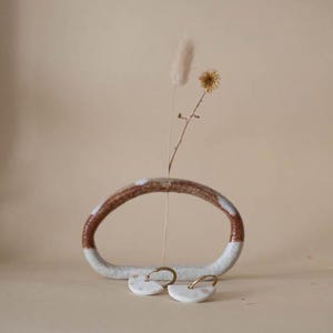 May include: A ceramic vase with a brown and white design, holding dried flowers. Two white and gold earrings are in front of the vase. The earrings have a half-moon shape.