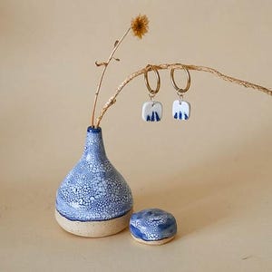 May include: A still life featuring a blue and white ceramic vase, a small matching dish, and a pair of gold hoop earrings with square, blue and white pendants. A dried flower stem is in the vase. The earrings and dish have a similar design.