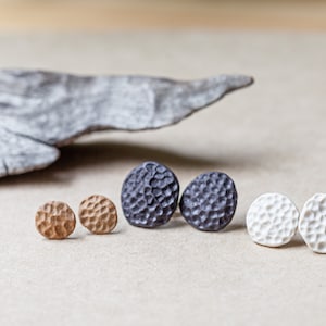 Op de afbeelding: Een verzameling handgemaakte oorstekers in verschillende kleuren. De oorbellen zijn in paren, met een textuur. Kleuren zijn bruin, donkergrijs en wit. Ze worden gepresenteerd op een neutrale ondergrond.