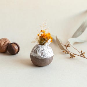Pode incluir: Um pequeno vaso redondo de cerâmica com a parte superior branca texturizada e a base castanha, contendo um pequeno buquê de flores secas laranja e brancas. O vaso é exibido em um fundo neutro, com nozes e folhas ao fundo.