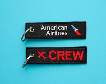 May include: Two black keychains with white and red embroidery. The top keychain says "American Airlines" with a red, white, and blue airplane logo. The bottom keychain says "CREW" with a red airplane logo.