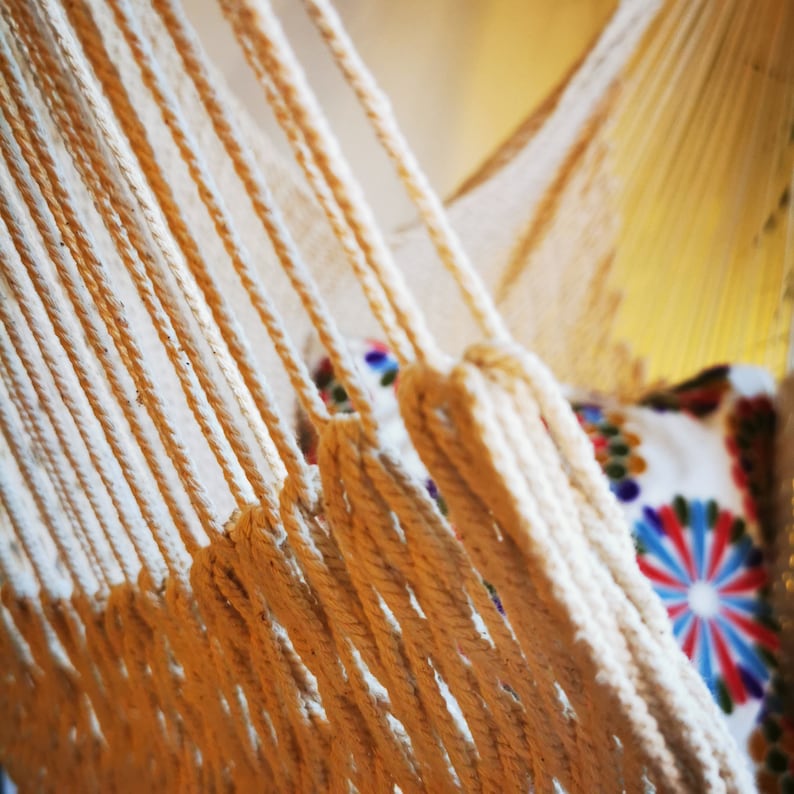 May include: Close-up of a white cotton hammock with thick, woven ropes. The hammock is hanging from a wooden frame and has a colorful patterned pillow.