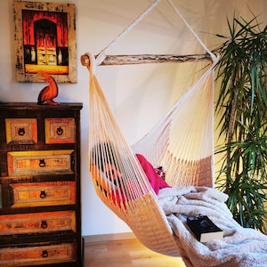 May include: A white hammock with a person relaxing inside. The hammock is hanging from a wooden beam and is made of a woven material. The person is wearing a red shirt and is lying down with a blanket over their legs. There is a green plant in the background.