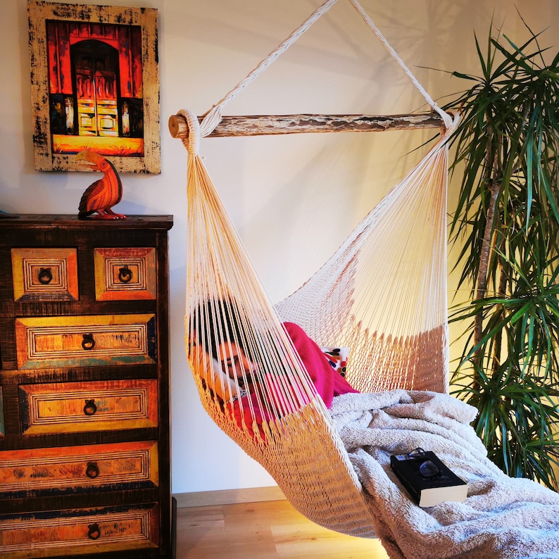 May include: A white hammock with a person relaxing inside. The hammock is hanging from a wooden beam and is made of a woven material. The person is wearing a red shirt and is lying down with a blanket over their legs. There is a green plant in the background.