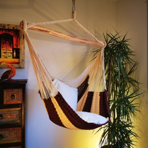 May include: A brown and beige hammock with a white cushion. The hammock is hanging from the ceiling and is in a room with a plant in the corner.