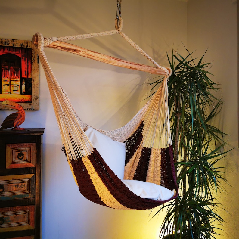 May include: A brown and beige hammock with a white cushion. The hammock is hanging from the ceiling and is in a room with a plant in the corner.