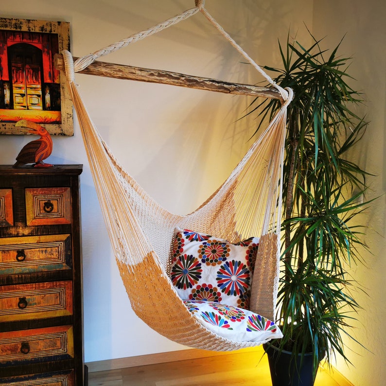 May include: A white hammock with a colorful pillow hanging from a wooden beam. The hammock is made of a woven material and has a fringe border. The pillow is decorated with a floral pattern.