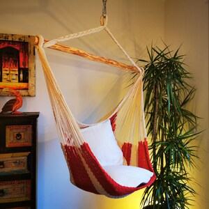 May include: A red and white hammock chair with a white cushion hanging from a wooden bar. The chair is in a room with a plant in the background.