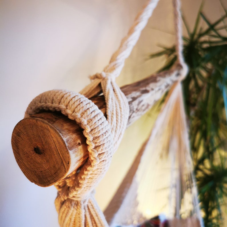May include: A white rope hammock with a wooden support beam. The hammock is hanging from the ceiling and is made of thick, natural rope. The wooden beam is attached to the rope with a knot.