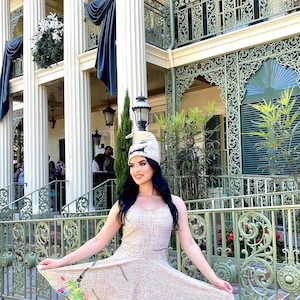 May include: A woman wearing a beige dress with a stitched pattern and a white hat with a stitched face. The dress has a fitted bodice and a flared skirt. The woman is standing in front of a building with a green metal railing.