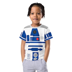 以下が含まれることがあります： 青と白のR2-D2のグラフィックプリントが施された白いTシャツ。シャツは半袖でクルーネックです。