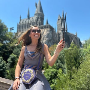 Könnte beinhalten: Eine Frau trägt ein graues Kleid mit einem blau-braun gestreiften Krawattenmuster und eine passende Handtasche mit dem Schriftzug "Chocolate Frog". Sie hält einen Zauberstab und trägt eine Sonnenbrille. Im Hintergrund ist ein großes Schloss.