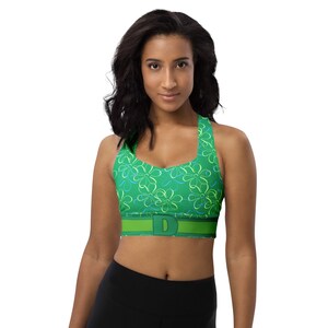 May include: Green sports bra with a floral pattern in shades of green and blue. The bra has a wide band with the letter "D" in the center. The model is wearing black leggings.