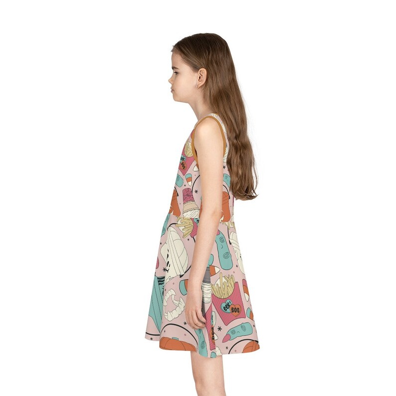 May include: A sleeveless dress with a light pink background and a colorful Halloween-themed pattern. The dress features a variety of cartoon illustrations, including a mummy, french fries, and the word "BOO". The dress has a brown trim.