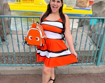 May include: A woman wearing an orange and white striped dress with a black and orange clownfish backpack and a matching headband. The background features a yellow submarine with the word "Nautilus" on the side.