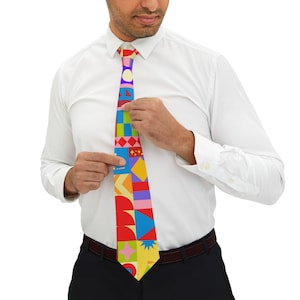 Op de afbeelding: Een kleurrijke stropdas met een geometrisch patroon van vormen in rood, geel, blauw, groen en paars. De stropdas wordt aangepast door een persoon die een wit overhemd en een zwarte broek draagt. Het ontwerp omvat vierkanten, driehoeken en cirkels.