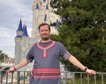 May include: A gray t-shirt with red and pink horizontal stripes and a red yoke design. The person is wearing khaki shorts and a black watch. The background features a large castle with blue spires and a cloudy sky.