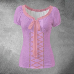 May include: A lilac-colored short-sleeved top with a pink ribbon and corset-style lacing. The top has a ruffled neckline and short, puffed sleeves. Gold-colored decorative accents are visible on the sides of the corset lacing.