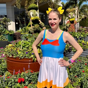 May include: A woman wearing a blue and white dress with a red bow and orange trim. She is standing in front of a flower garden with topiary sculptures of Mickey Mouse and Donald Duck.