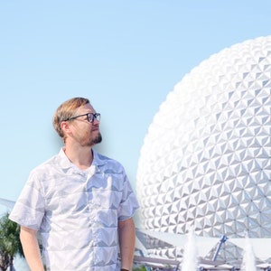 Puede incluir: Una camisa de manga corta con estampado geométrico en gris claro y blanco. La camisa se usa con jeans azules y un reloj marrón. El fondo presenta una gran esfera geodésica blanca y una fuente bajo un cielo azul claro.