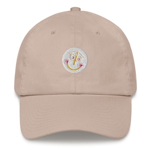 Puede incluir: Una gorra de béisbol beige con una cara sonriente blanca bordada con una cruz amarilla y fuegos artificiales rosas.