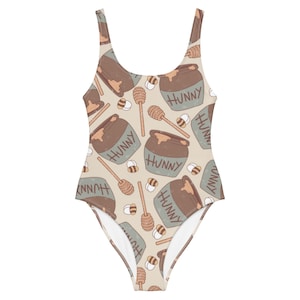 May include: One-piece swimsuit with a repeating pattern of honey pots, honey dippers, and bees. The honey pots are brown with the word "HUNNY" printed on them. The swimsuit has a light tan background.
