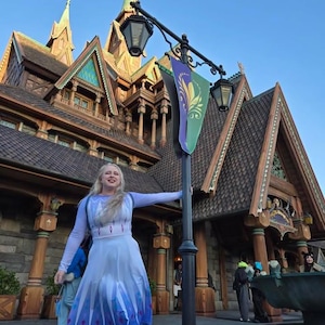 Pode incluir: Uma mulher vestida como Elsa de Frozen está em frente a um prédio de madeira com uma bandeira verde e roxa. Ela está segurando um poste de luz com a mão direita e sorrindo.