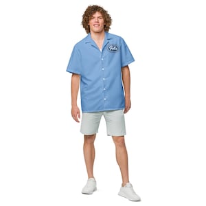 Pode incluir: Uma camisa azul claro de manga curta com colarinho branco e um remendo oval azul que diz "Felix". A camisa está sendo usada por uma pessoa com cabelo castanho encaracolado. A pessoa está usando shorts cinza claro e tênis brancos.