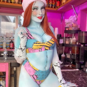 Puede incluir: Una mujer con un bikini de patchwork azul y rosa, un gorro de Papá Noel blanco y guantes blancos. Tiene pintura facial azul y blanca con líneas cosidas y labios rojos. Está de pie frente a un mostrador rosa y blanco.