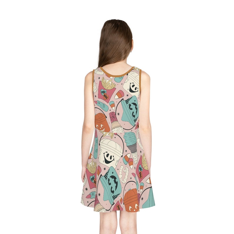 May include: A sleeveless dress featuring a repeating pattern of Halloween-themed illustrations. The design includes smiling mummy faces, ice cream cones, french fries, and candy corn on a light pink background. The dress has a brown trim.