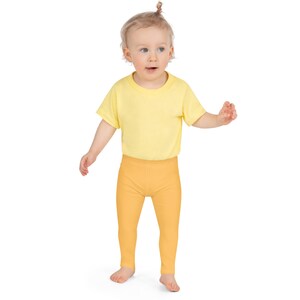 May include: A toddler wearing a yellow t-shirt and solid orange leggings. The child is standing and looking to the side. The outfit is simple and comfortable, suitable for everyday wear.