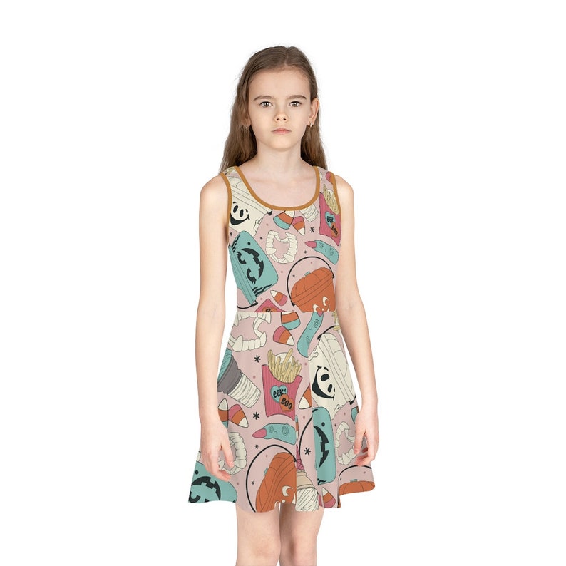 May include: A sleeveless dress with a pink background and a repeating pattern of Halloween-themed illustrations. The pattern includes images of ghosts, pumpkins, french fries, and candy corn. The dress has a brown trim.
