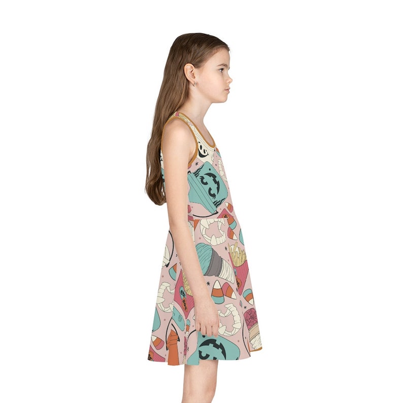 May include: A sleeveless dress featuring a Halloween-themed pattern. The dress has a light pink background with various Halloween-related illustrations in teal, orange, and white. The dress is worn by a person.