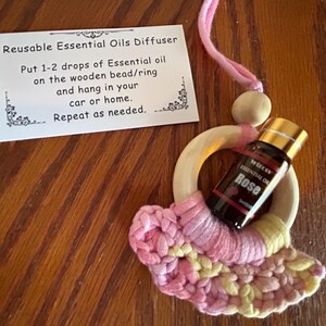 May include: A reusable essential oil diffuser with a wooden ring and a pink and yellow crocheted base. A small bottle of rose essential oil is nestled within the ring. A card provides instructions for use.